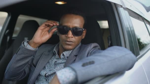 Retrato de un hombre de negocios serio sentado dentro del coche puesto de gafas de sol y sonriendo en la cámara al aire libre — Vídeos de Stock