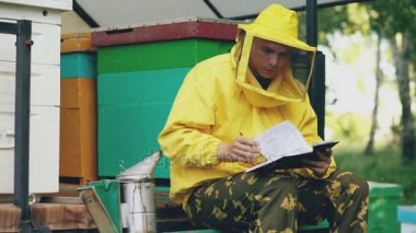 Genç arıcı adam yazmak notepad arı kovanı yanında otururken hasat kontrol