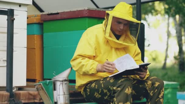 Young beekeeper man write in notepad checking harvest while sitting near beehives — Stock Video