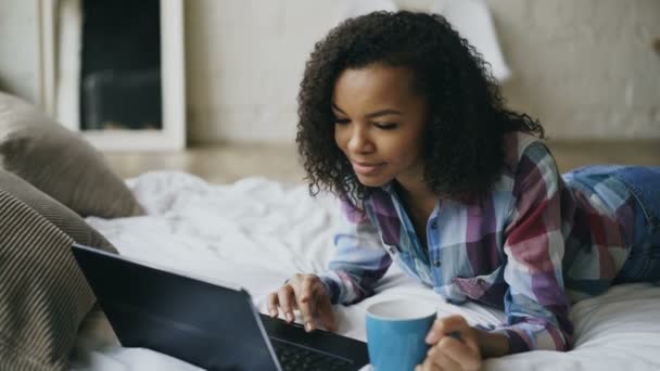 Jovem menina africana atraente usando laptop para compartilhar mídia social deitada na cama em casa — Vídeo de Stock