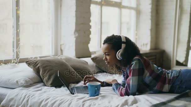 Atraktivní smíšený závod dívka poslouchat hudbu při procházení sociální média na notebooku ležící na posteli — Stock video