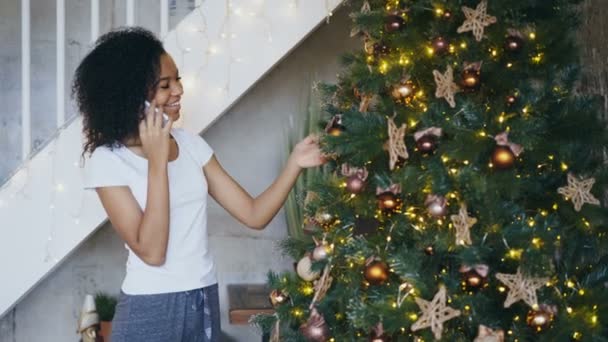 Chica de raza mixta rizada decorando el árbol de Navidad en casa y charlando con el teléfono inteligente — Vídeos de Stock