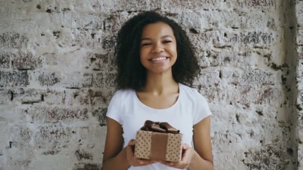 Retrato de la joven chica africana feliz sosteniendo caja de regalo y sonriendo en la cámara — Vídeos de Stock