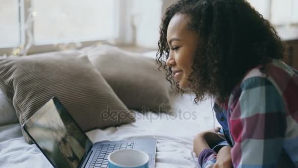 Alegre misto raça menina ter vídeo chat com amigos usando laptop câmera enquanto deitado na cama — Vídeo de Stock