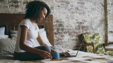 Evde yatakta oturmuş sosyal medya paylaşımı için dizüstü bilgisayarda yazarak neşeli karışık yarış kız