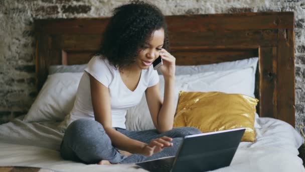 Curly menina americana africana falando smartphone e usando computador portátil sentado na cama em casa — Vídeo de Stock