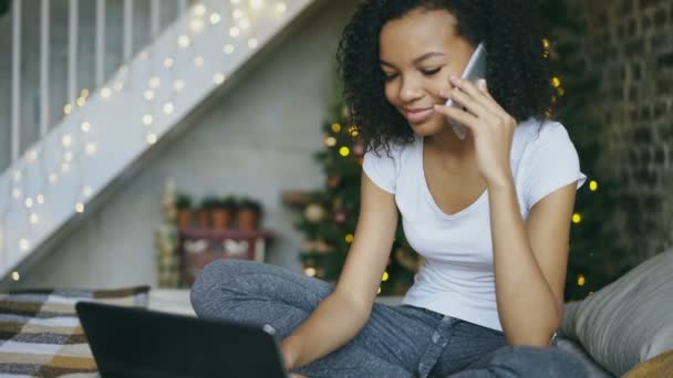 Krullend gemengd ras meisje chatten smartphone en laptopcomputer via kerstboom backround thuis — Stockvideo