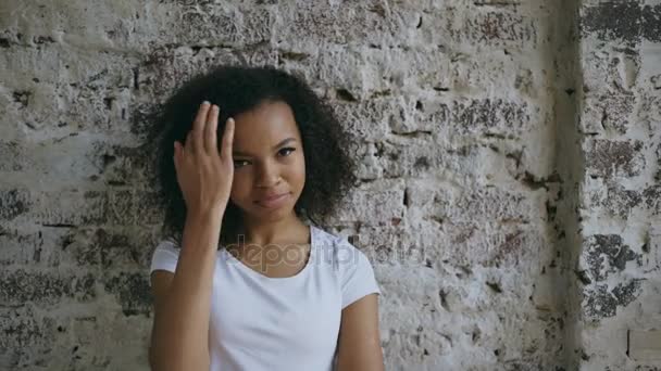 Портрет молодої усміхненої змішаної раси жінки фліртує і дивиться в камеру на фоні цегляної стіни — стокове відео