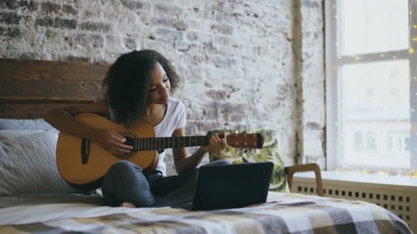 Concentraing dziewczyna kręcone Afryki amerykański nastolatek nauka gry na gitarze przy użyciu komputera przenośnego, siedząc na łóżku w domu — Wideo stockowe