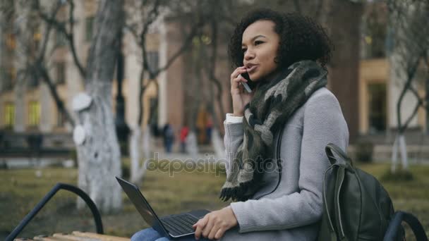 Attractive african american student girl talking her phone sitting on bench with laptop computer outdoors — Stock Video