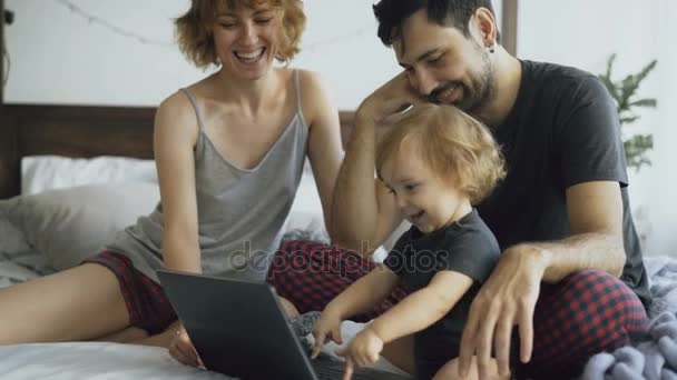 Família jovem com linda menina brincando com a filha enquanto ela digitando no computador portátil sentado na cama em casa — Vídeo de Stock