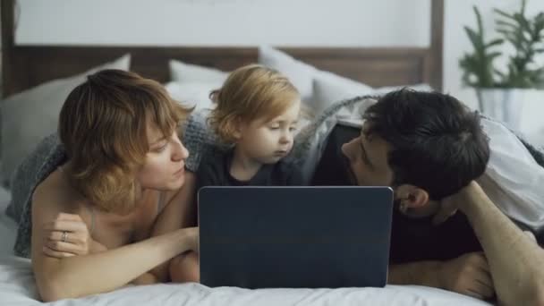 Familia joven con una linda niña jugando con su hija mientras ve la computadora portátil tumbada en la cama en casa — Vídeo de stock