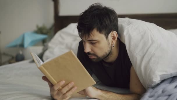 Красивый молодой человек читает книгу, лежащую в постели под одеялом дома — стоковое видео