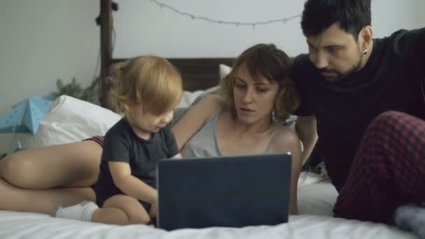 Família jovem com linda menina brincando com a filha enquanto ela digitando no computador portátil sentado na cama em casa — Vídeo de Stock