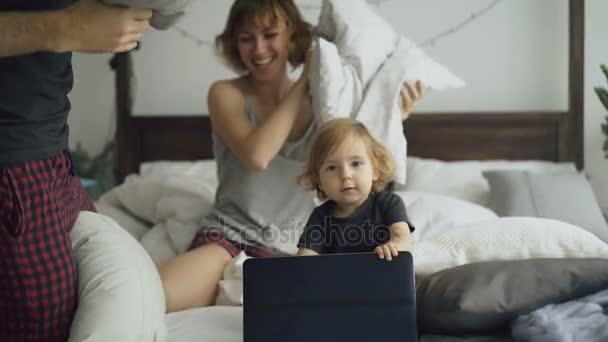 Счастливая семья с маленькой симпатичной дочкой, играющей и сражающейся подушками в постели дома — стоковое видео