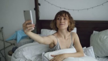 Neşeli kadın evde yatakta otururken smartphone kullanarak selfie portre çekici
