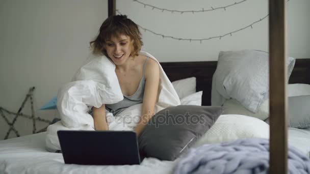 Joven alegre usando su computadora portátil sentada en la cama en casa — Vídeo de stock