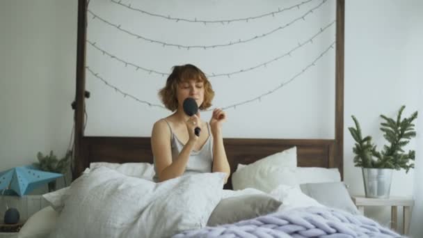 Atractiva mujer alegre bailando y cantando con peine como micrófono sentado en la cama en casa — Vídeos de Stock