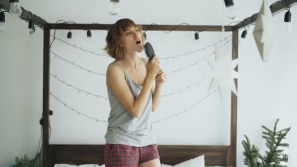 Atraktivní žena veselý tanec na posteli a zpěv s hřeben jako mikrofon doma — Stock video