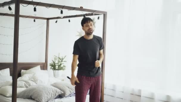 Молодой привлекательный мужчина танцует и поет как рок-музыкант у постели дома — стоковое видео