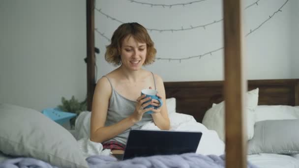 Atractiva joven mujer que tiene chat de vídeo en línea con amigos utilizando la cámara portátil mientras está sentado en la cama en casa — Vídeo de stock