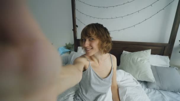 Vrolijke jongedame portretje selfie met smartphone zitten in bed in thuis — Stockvideo