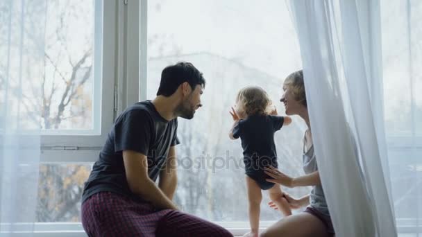 Familia feliz con joven linda hija sentada en el alféizar de la ventana jugando y mirando en la ventana en casa — Vídeos de Stock