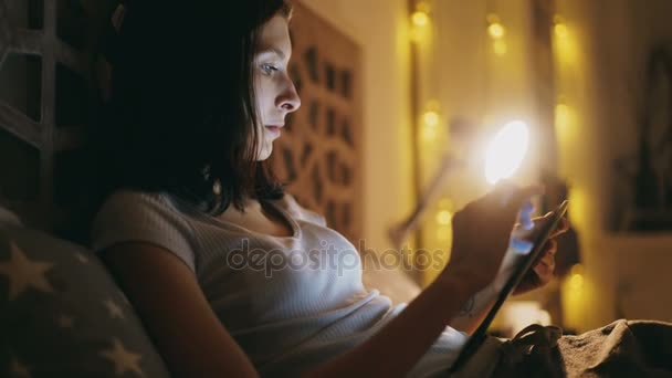 Primo piano di una giovane donna concentrata che usa tablet e guarda le notizie online sdraiata a letto di notte — Video Stock