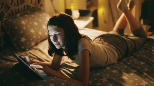 Atractiva mujer sonriente relajándose en la cama usando tableta por la noche en casa — Vídeo de stock