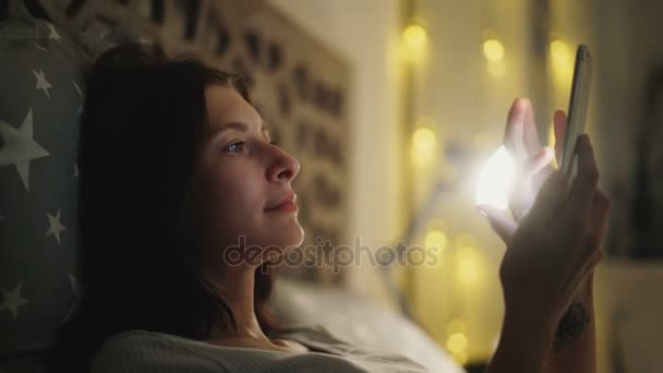 夜、自宅のベッドに横になっている彼女のスマート フォンでオンライン ゲームを遊んで笑顔の女性 — ストック動画