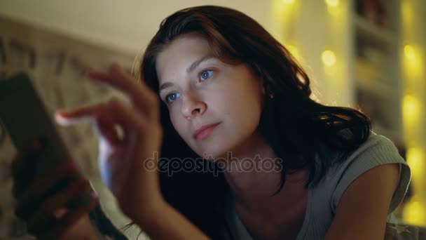 Primer plano de la joven mujer concentrada utilizando el teléfono inteligente acostado en la cama en casa por la noche — Vídeo de stock