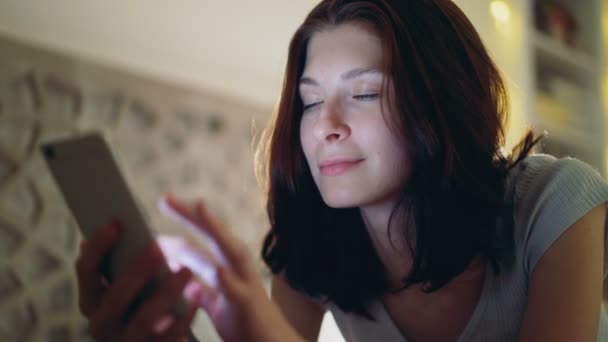 Nahaufnahme einer jungen lächelnden Frau mit dem Smartphone, die nachts zu Hause im Bett liegt — Stockvideo