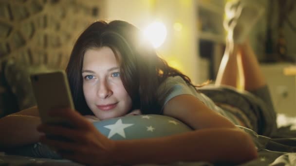 밤에 집에서 침대에 누워 하는 소셜 미디어를 공유 하기 위한 스마트폰을 사용 하 여 매력적인 웃는 여자 — 비디오
