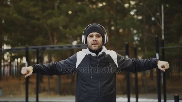 Красивый человек в наушниках делает разминку, слушая музыку в зимнем парке — стоковое видео