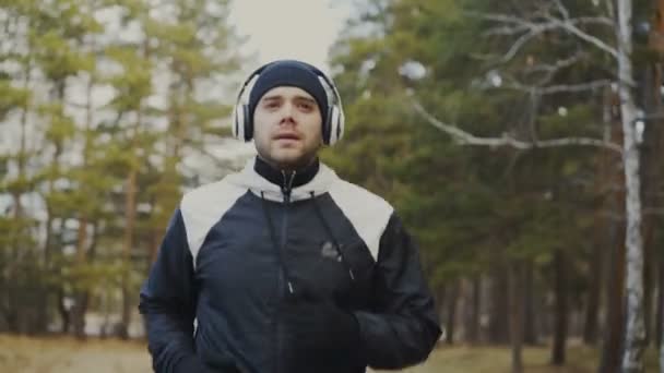 冬の公園で音楽を聴く中朝のジョギングのヘッドフォンでハンサムなランナーの人のトラッキング ショット — ストック動画