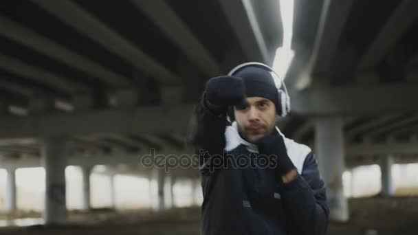 Tracking shot de Joven boxeador en auriculares entrenamiento golpes en la ubicación urbana al aire libre en invierno — Vídeos de Stock