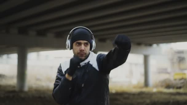 Стеження за зйомками спортивного боксера в навушниках, що роблять вправи з боксу в міському місці на відкритому повітрі взимку — стокове відео