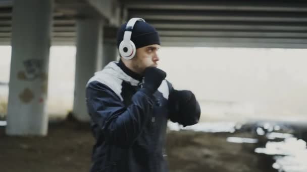 Rastreamento tiro de homem esportivo boxeador em fones de ouvido fazendo exercício de boxe em localização urbana ao ar livre no inverno — Vídeo de Stock