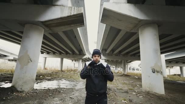 Jovem boxeador em fones de ouvido fazendo exercício de boxe enquanto ouve música em localização urbana ao ar livre no inverno — Vídeo de Stock