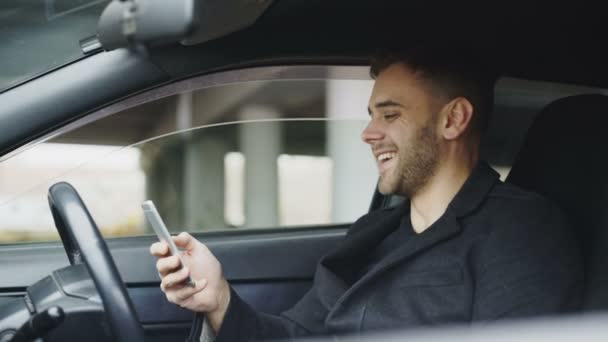 Empresário atraente sentado dentro do carro rindo enquanto usa smartphone após viagem — Vídeo de Stock