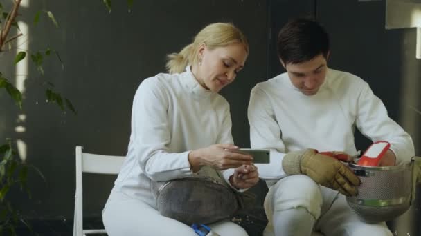 Two young fencers man and woman watching fencing competition on smartphone and sharing experience after training indoors — Stock Video