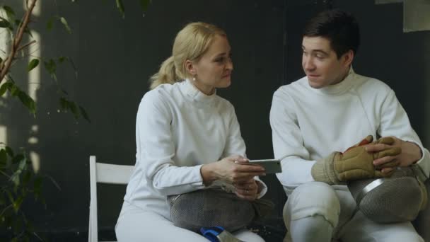 Two young fencers man and woman watching fencing competition on smartphone and sharing experience after training indoors — Stock Video