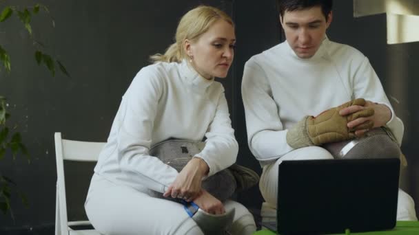 Двоє молодих фехтувальниць чоловік і жінка спостерігають за фехтуванням на портативному комп'ютері і обмінюються досвідом перед тренуванням в приміщенні — стокове відео
