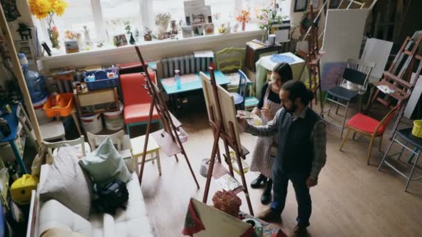 Geschoolde kunstenaar man onderwijs jonge vrouw schilderij op ezel op school van kunststudio - creativiteit, onderwijs en kunst mensen concept — Stockvideo
