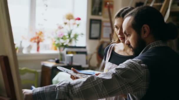 Geschoolde kunstenaar man onderwijs jonge vrouw schilderij op ezel op school van kunststudio - creativiteit, onderwijs en kunst mensen concept — Stockvideo