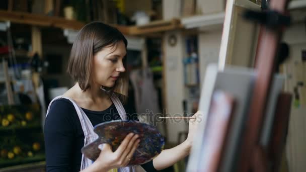 アート スタジオでキャンバスに絵を描いているエプロンの若い女性アーティスト — ストック動画