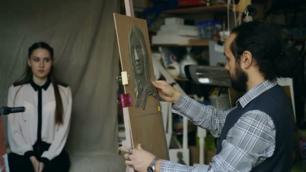 Скульптор создает скульптуру человеческого лица на холсте, пока молодая женщина позирует ему в художественной студии — стоковое видео