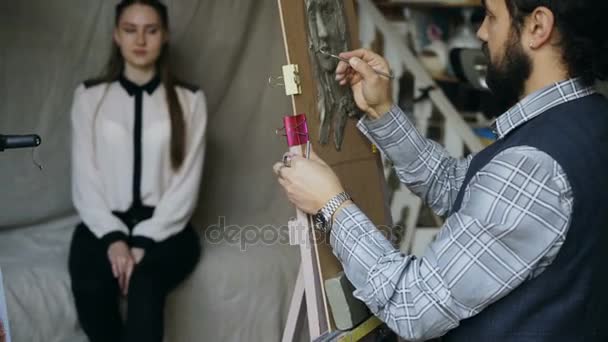 Sculptor creating sculpture of humans face on canvas while young woman posing him in art studio — Stock Video