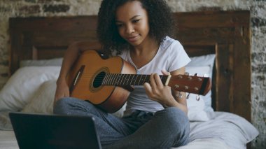 Evde yatakta otururken dizüstü bilgisayar kullanan gitar çalmayı öğrenme çekici Afrikalı-Amerikalı genç kız concentraing