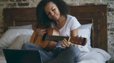 Evde yatakta otururken dizüstü bilgisayar kullanan gitar çalmayı öğrenme çekici Afrikalı-Amerikalı genç kız concentraing